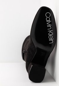 Bottine en daim noir à talon bloc, semelle tournée vers l'avant montrant le logo « Calvin Klein » en lettres blanches.