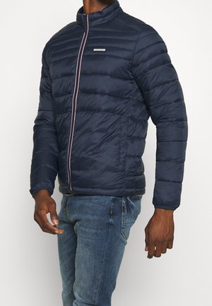 Mand iført en marineblå quiltet lynlåsjakke og blå jeans, stående mod en ensfarvet lys baggrund.