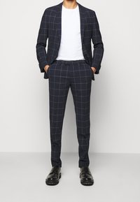 Sininen ruudullinen puku, jossa on slim fit -leikkaus ja joka koostuu takista ja yhteensopivista housuista. Kangas on sileä. Mustat kengät täydentävät asukokonaisuutta.
