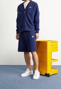 Marinblå Nike cardigan med knappar, kombinerad med matchande marinvita shorts. Vit Nike-logga på båda delarna. Vita sneakers och en gul kundvagn i närheten.