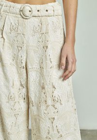 Pantaloni in pizzo color crema con intricati motivi floreali, una cintura larga e una cintura rotonda con un singolo bottone dorato.