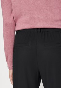 Calças pretas caneladas com um cós elástico e dois bolsos traseiros, combinadas com um top de malha de mangas longas em rosa claro.