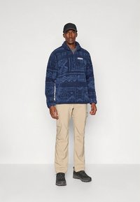 Sötétkék polár pulóver texturált mintával és félig cipzáros kialakítással, bézs cargo nadrággal és fekete túracipővel, egy férfi által viselve.