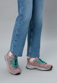 Jasnoróżowe buty sportowe z turkusowymi sznurowadłami, teksturowanymi panelami i szarymi podeszwami, zestawione z niebieskimi dżinsami i białymi skarpetkami.