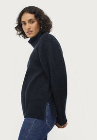 Marineblauwe coltrui met een geribbelde textuur, zijsplitten en een ontspannen pasvorm. Gepaard met donkerblauwe jeans, weergegeven vanuit een zijaanzicht.