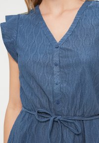 Μπλε φόρεμα με υφή που διαθέτει V-λαιμόκοψη, κοντά φουσκωτά μανίκια, κουμπιά μπροστά και λεπτομέρεια με δεμένη μέση. Η επιφάνειά του έχει ανάγλυφο μοτίβο κυματισμών.