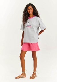 Šedé oversized tričko s textem "CHODÍM SE PSEM, TO JE MÉ KARDIO" a designovým detailem. Kombinováno s růžovými lněnými šortkami a hnědými sandály na řemínky.