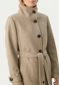 Cappotto in lana beige con colletto alto, quattro bottoni neri e cintura. Presenta tasche laterali e polsini ripiegati per un tocco di dettaglio in più.