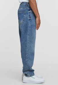 Jeans in denim azzurro chiaro con una vestibilità rilassata, caratterizzati da una vita alta, tasche laterali e una texture leggermente sbiadita. Indossati con sneakers bianche.