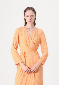 Rochie texturată portocalie, cu mâneci lungi și decolteu în V, având un cordon în față și un model de carouri discret. Material neted.