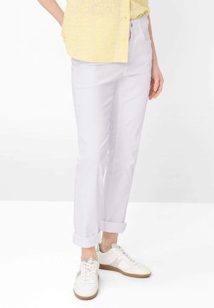 Persona che indossa pantaloni bianchi risvoltati, sneakers bianche con dettagli argentati e una camicia gialla a maniche corte con bottoni, in piedi su uno sfondo bianco.