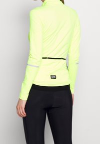 Pitkähihainen, kirkkaan neonkeltaisesta materiaalista valmistettu juoksupaita, jossa on vetoketjullinen tasku, musta vyötärö ja heijastavat yksityiskohdat hihoissa. Yhdistetty mustiin legginsseihin.