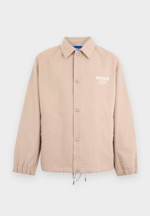 Bézs gombos dzseki gallérral, elasztikus mandzsettával, megkötős alsó résszel és fehér "HUGO" logóval a bal mellrészen.