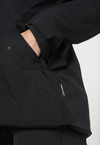 Black jacket made of textured fabric, featuring a fitted design, side pocket with snap closure, and a small branded label at the hem.