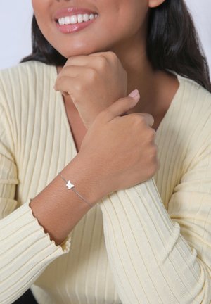 Sølvarmbånd med en lille hvid sommerfuglecharms, båret på et håndled. Bæreren har en lys gul ribbet sweater og en brun hudfarve.