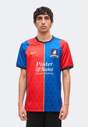 Jeune homme portant un maillot de football divisé verticalement rouge et bleu avec un logo Nike jaune et un texte de sponsor blanc à l'avant.