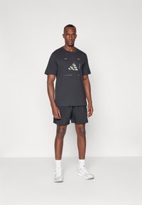 Camiseta negra de manga corta con un diseño gráfico, combinada con pantalones cortos negros. El modelo lleva calcetines blancos y zapatillas deportivas gris claro.
