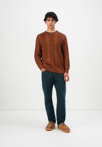 Giovane in piedi, con un maglione marrone a trecce, pantaloni verde scuro e scarpe scamosciate beige su uno sfondo bianco semplice.
