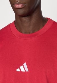 Sarkans kokvilnas T-krekls ar apaļu kakla izgriezumu un īsām piedurknēm. Kreisajā krūšu pusē redzams balts Adidas logotipa attēls.