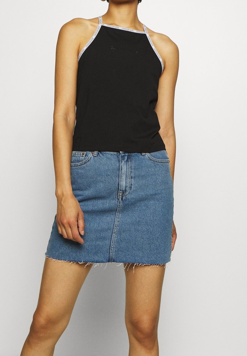 Kvinna i svart halterneck-topp med vit kant och blå denimkjol med fransad nederkant, står mot en enfärgad ljus bakgrund.