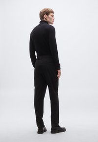 Čierna polokošeľa s dlhým rukávom a vysokým golierom v kombinácii s čiernymi elegantnými nohavicami na mieru. Model stojí otočený chrbtom, obuv má hladký povrch.
