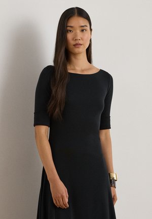 Femme aux longs cheveux foncés portant une robe noire à manches jusqu'au coude et un grand bracelet doré et marron à son poignet gauche.