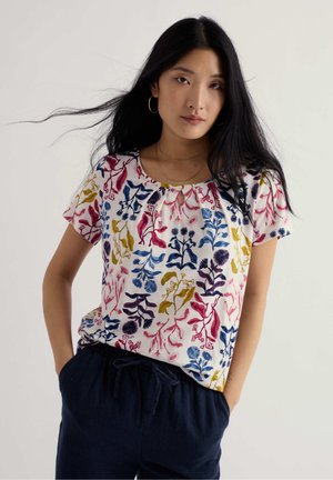 Femeie cu păr lung și negru poartă un top alb cu mâneci scurte și imprimeuri florale multicolore, precum și pantaloni închiși la culoare cu șnur, pozând cu mâinile în buzunare.