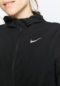 Gros plan sur une personne portant une veste noire zippée avec capuche et un logo blanc Nike sur la poitrine.