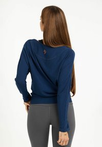 Marineblaues Langarmoberteil mit entspannter Passform, Rundhalsausschnitt und orangefarbenem gesticktem Logo auf dem Rücken, kombiniert mit grauen engen Leggings.