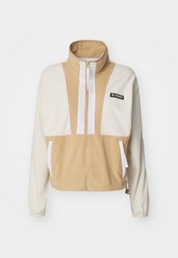 Beskuren fleecejacka med hög krage, i beiga och krämfärgade block, vit dragkedja och två framfickor. Columbia-logotyp nära dragkedjan.