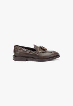 Mocasin de cuero marrón con punta redonda, borlas decorativas, costuras en contraste y una suela de bajo perfil. Textura suave con un acabado pulido.