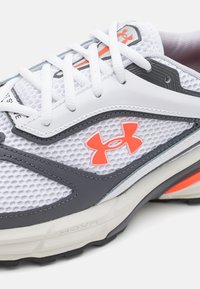 White mesh athletic shoe with grey accents, featuring a prominent orange logo, textured surface, and white laces. Lightweight construction.