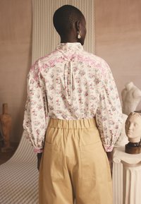 Persoană purtând o bluză cu mâneci lungi cu imprimeu floral și pantaloni bej cu talie înaltă, stând cu spatele spre cameră într-o cameră cu sculpturi.