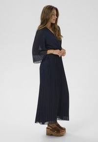 Marineblaues, plissiertes Maxikleid mit weiten Glockenärmeln, V-Ausschnitt und strukturierter Stoffstruktur. Kombiniert mit braunen Kork-Plateausandalen.