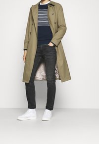 Beige trenchcoat över en marinblå tröja med geometriskt mönster, matchat med mörka jeans och vita sneakers. Slät tyg och skräddarsydd passform.