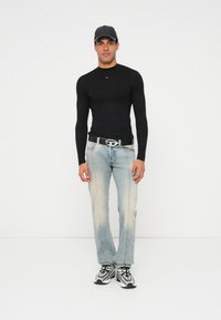 Schwarzes Langarmshirt, hellblaue, ausgewaschene Jeans, Gürtel mit silberner Schnalle, schwarze Mütze und weiß-silberne Turnschuhe, stehend vor einem schlichten Hintergrund.