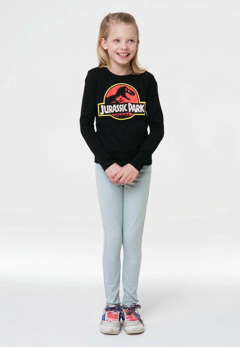 Schwarz langärmliges Shirt mit einem roten und gelben "Jurassic Park"-Logo, kombiniert mit hellblauen Hosen und mehrfarbigen Turnschuhen.