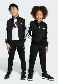 Černé sportovní teplákové soupravy s bílými pruhy, obsahující zapínací bundy a odpovídající kalhoty. Děti nosí černé tenisky s bílými akcenty.