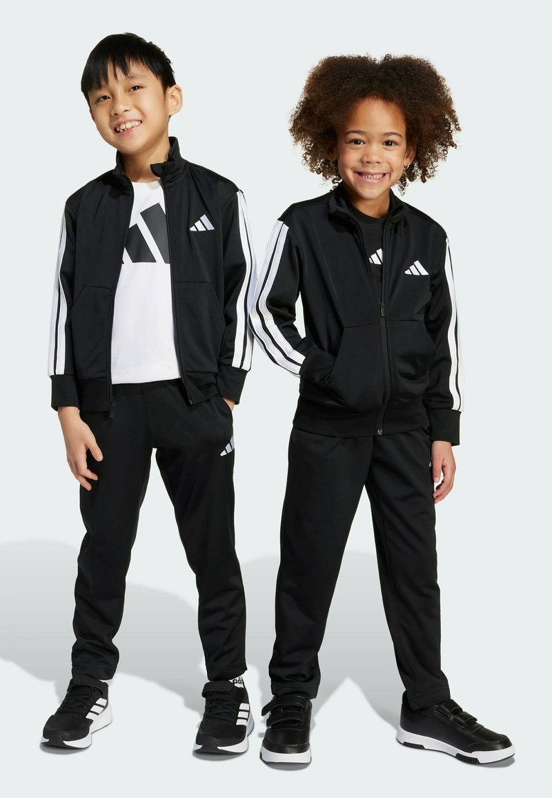 Černé sportovní teplákové soupravy s bílými pruhy, obsahující zapínací bundy a odpovídající kalhoty. Děti nosí černé tenisky s bílými akcenty.