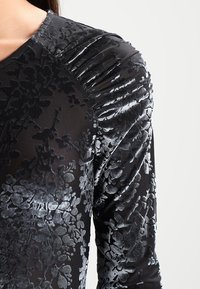 Svart långärmad topp med blommig sammetstextur, med rynkade detaljer på axlarna och en slät tygkropp.