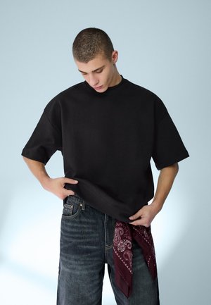 Jeune homme portant un t-shirt noir oversize et un jean foncé, ajustant un bandana bordeaux à motif suspendu à sa poche, sur un fond uni.