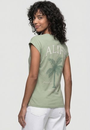 Frau mit lockigem Haar, die ein hellgrünes T-Shirt mit einer Palme und dem Schriftzug "ALIFE" trägt, leicht gedreht und lächelt über ihre Schulter.