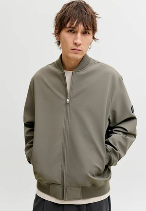 Ung person iført en grå bomberjakke med lynlås og hænderne i lommerne, stående mod en ensfarvet lys baggrund.