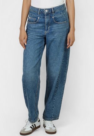 Person trägt locker sitzende blaue Jeans, weiße Turnschuhe mit schwarzen Streifen und ein weißes Oberteil, steht vor einem schlichten Hintergrund.