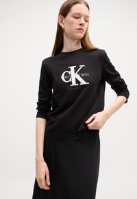 Juoda ilga rankovė marškinėliai su balta "CK" logotipo ir teksto dizainu priekyje, pagaminti iš švelnios medvilnės medžiagos, su laisvu siluetu.