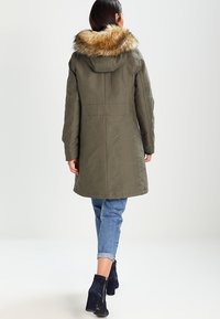 Parka verde oliva con capucha de piel sintética, con un corte recto, textura acolchada y detalles en la costura trasera. Combinada con jeans arremangados.