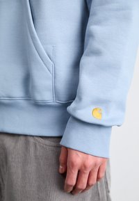 Pulover bleu deschis, cu mansete și tiv cu nervuri, având un logo galben pe mânecă. Materialul pare moale și texturat.