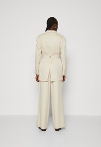 Beige Leinenanzug mit taillierter Jacke und weit geschnittenen Hosen. Verfügt über eine taillierte Taille und einen Rückenschlitz, kombiniert mit schwarzen Absätzen.