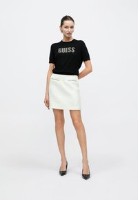 Schwarzes kurzärmeliges Oberteil mit „GUESS“ in metallischem Schriftzug, kombiniert mit einem weißen, strukturierten Minirock. Model trägt schwarze High Heels.