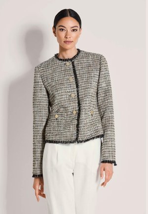 Frau mit zurückgebundenem dunklem Haar trägt eine strukturierte karierte Jacke mit goldenen Knöpfen und schwarzem Besatz, kombiniert mit weißen Hosen vor hellem Hintergrund.